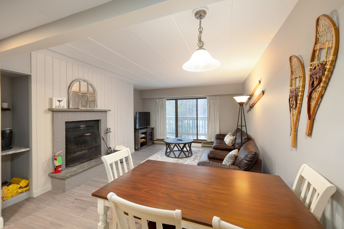An open living area features a wooden dining table with white chairs near a cozy seating arrangement. The fireplace is positioned centrally, and large windows lead to a balcony, allowing natural light to fill the space. Decorative snowshoes are displayed on the wall.