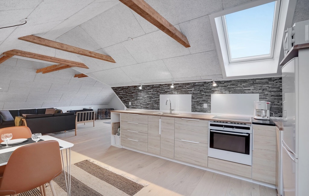 An open kitchen and living area feature modern wooden cabinetry and an oven. Large windows allow natural light to fill the space, highlighting the textured stone wall. A cozy seating area with a sofa and dining table is visible, offering a functional living experience.
