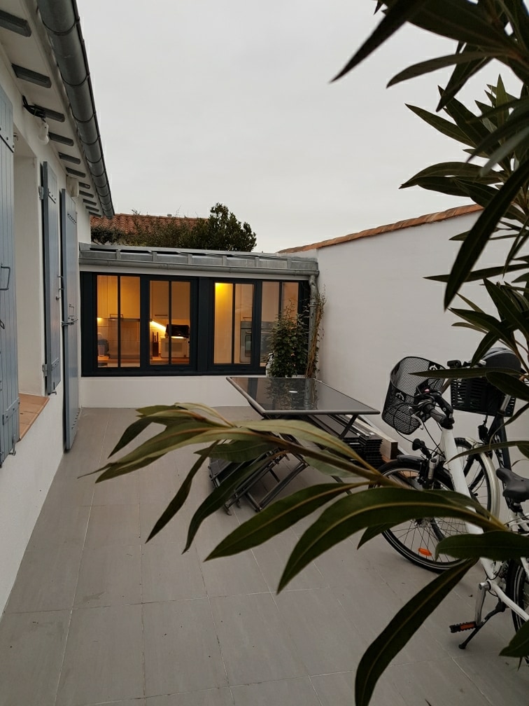 A terrace area is visible, featuring a glass table surrounded by chairs. Lush greenery is present in pots, and a bicycle is parked nearby. The entrance to the interior space is accessible through large glass doors, inviting natural light into the area.