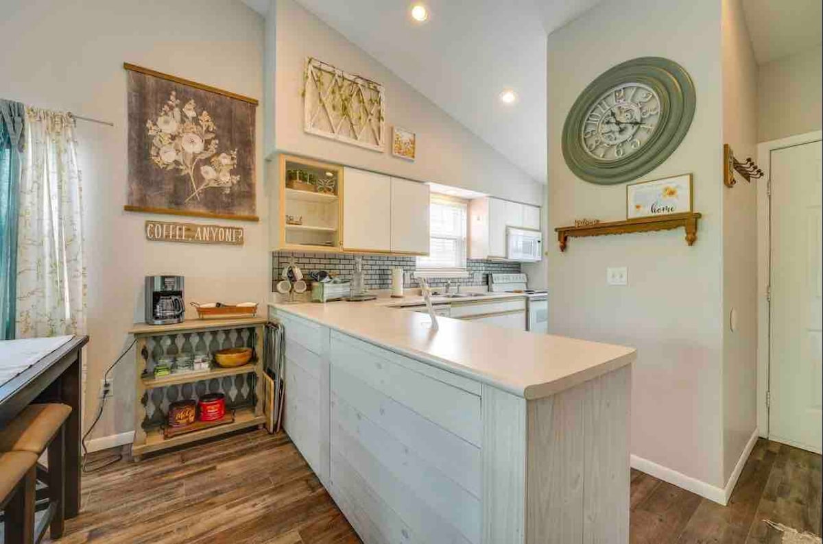 A modern kitchen is displayed with white cabinets and a light countertop. A coffee station is set up beside a wall clock, while decorative wall art adds character. Natural light is filtered through sheer curtains, highlighting the spacious layout and accessible design.