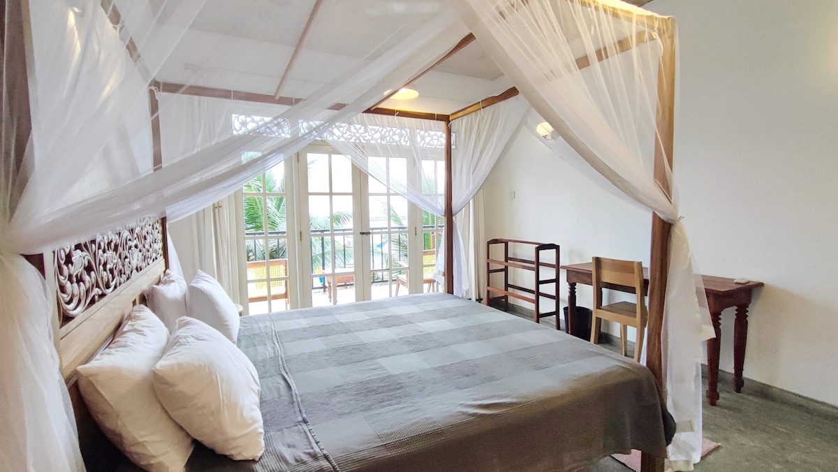 A spacious room features a large bed draped with delicate sheer fabric, enhancing the airy feel. Natural light streams through French doors leading to a private balcony, providing a view of palm trees. A writing desk and an open wardrobe offer practical furnishings for guests.