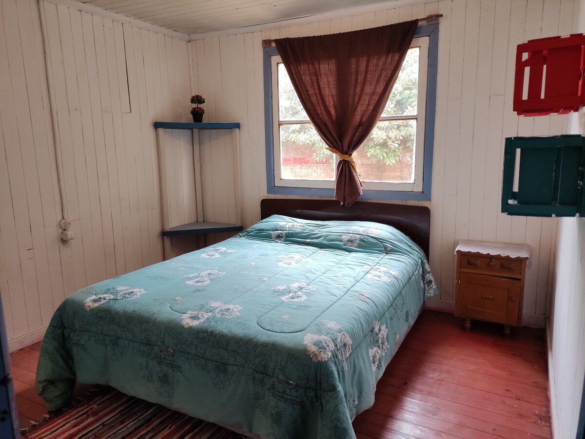 The private room features a spacious bed covered with a light blue quilt. Natural light enters through a large window adorned with a dark curtain. A small nightstand and open shelving add functionality to the room, which is accented by wooden flooring.