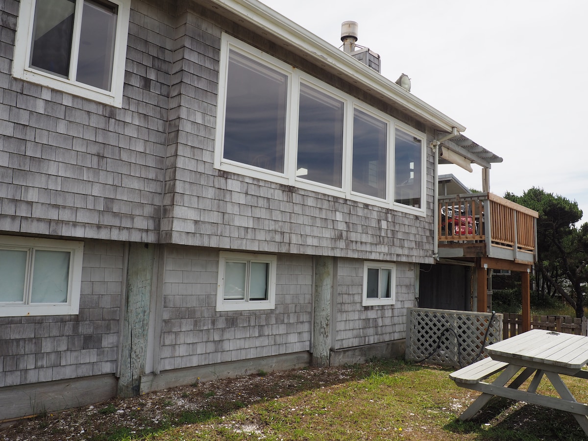 The exterior of a coastal home is shown, featuring a mix of weathered wooden shingles and large windows that provide natural light. A small deck with seating is visible, along with a picnic table situated on the grass, creating a casual outdoor space.