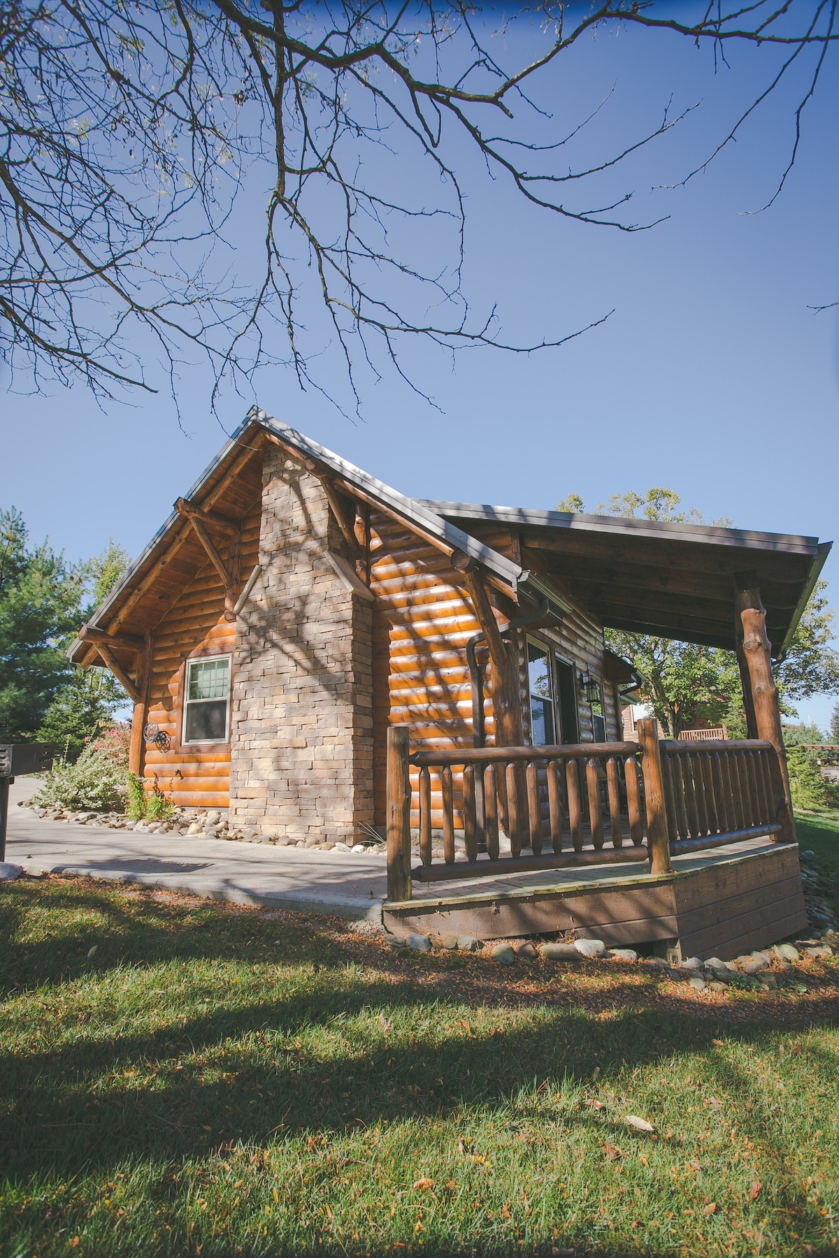 The standalone cabin features rustic log and stone construction, complemented by an inviting front porch with wooden railing. Surrounding grassy areas enhance the serene atmosphere, while clear blue skies provide a bright backdrop to this charming retreat.