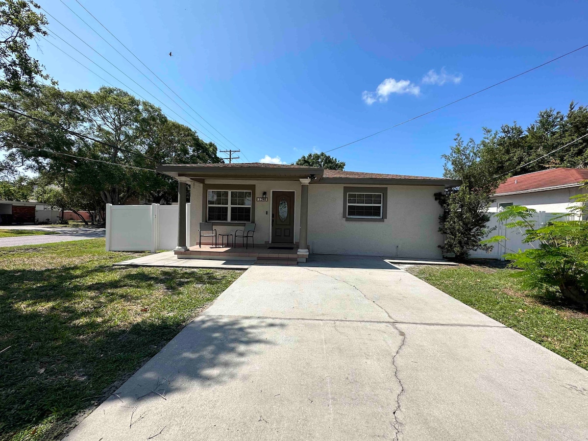 A single-story home is featured with a welcoming porch and a simple front entrance. A small seating area with two chairs is positioned on the porch. The driveway leads to a clear, open area, surrounded by green grass and a few trees enhancing the exterior setting.
