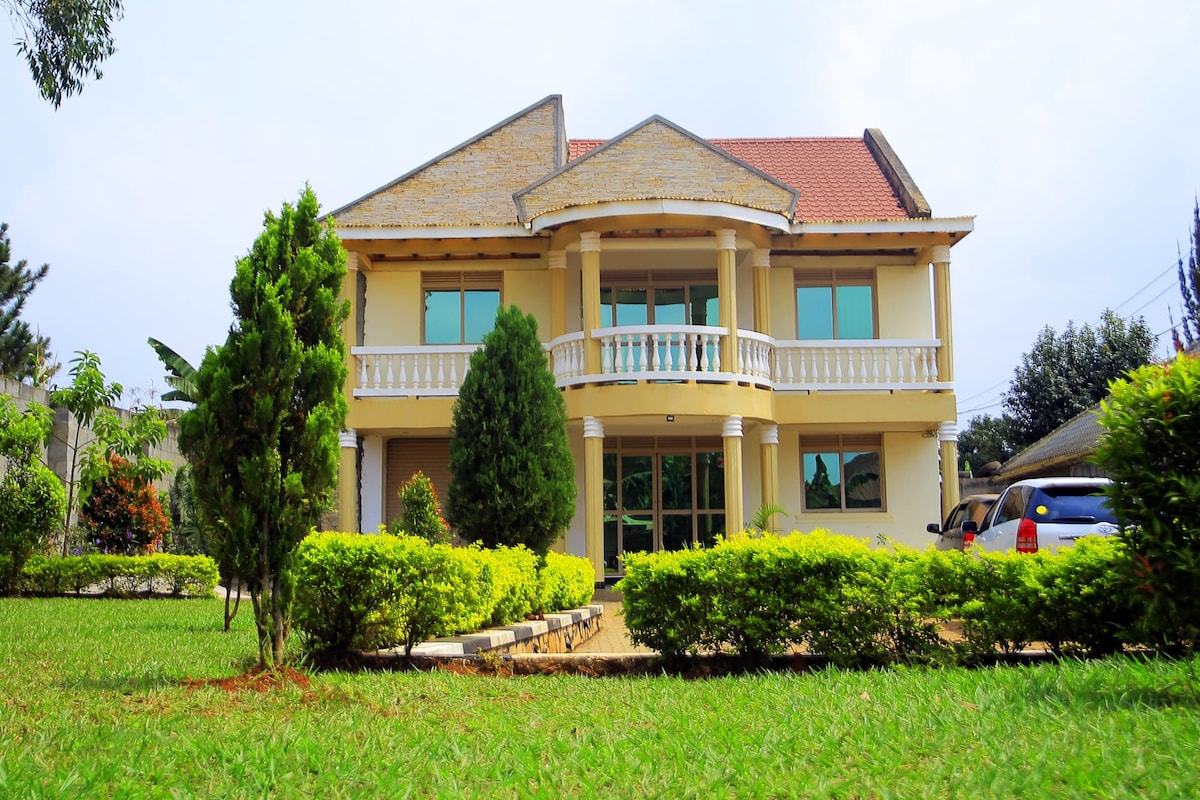 A well-maintained three-story building features a welcoming entrance with a large balcony. Lush green shrubs and trees surround the property, complemented by a neatly manicured lawn. The driveway leads towards a parked car, emphasizing ease of access.