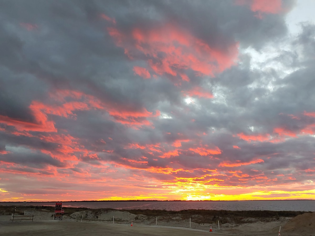 A vibrant sky is filled with swirling clouds in shades of gray, orange, and pink, creating a stunning sunset over a coastal landscape. The horizon glows warmly, meeting a vast expanse of sand and water, suggesting a serene and picturesque evening.