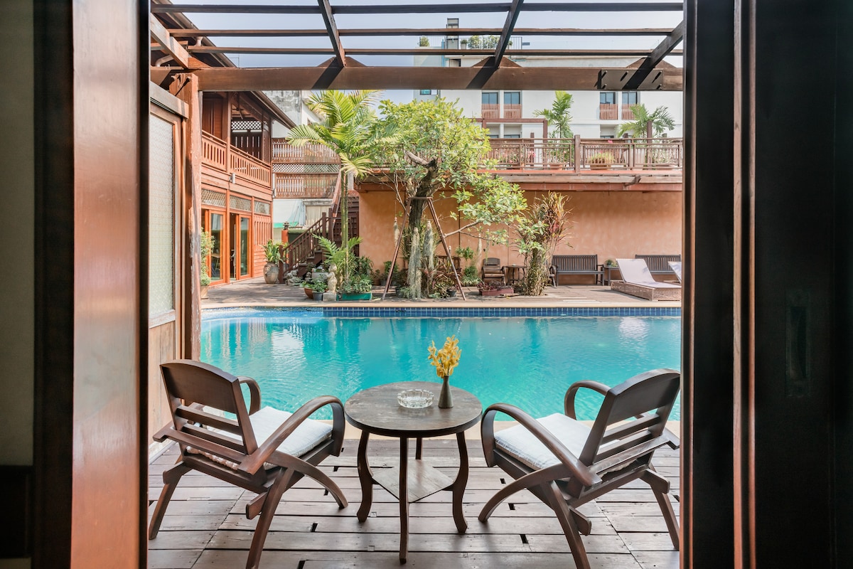 A private patio features two wooden chairs and a round table, facing a serene outdoor saltwater pool. Lush greenery frames the view, and the tranquil water reflects the surrounding architecture, inviting relaxation and social gatherings.