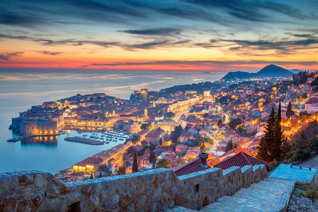 A panoramic view showcases the illuminated city of Dubrovnik during twilight, with subtle hues of orange and blue in the sky. The coastline features a harbor filled with boats, while stone walls and historical buildings are visible throughout the landscape, giving a sense of the city's rich history.