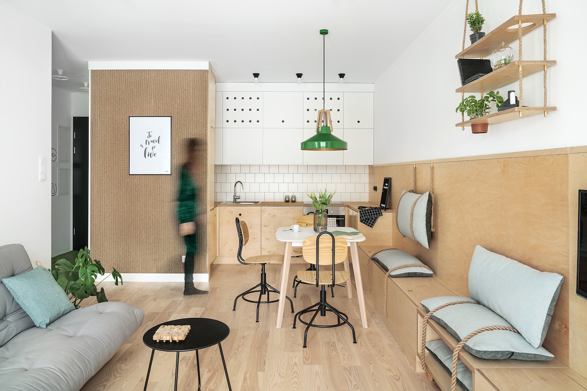 A modern living space features a blend of wood and greenery. A round dining table with black chairs sits in the center, while a cozy, gray sofa offers additional seating. The kitchen area is designed with sleek cabinetry and is illuminated by a green pendant light.