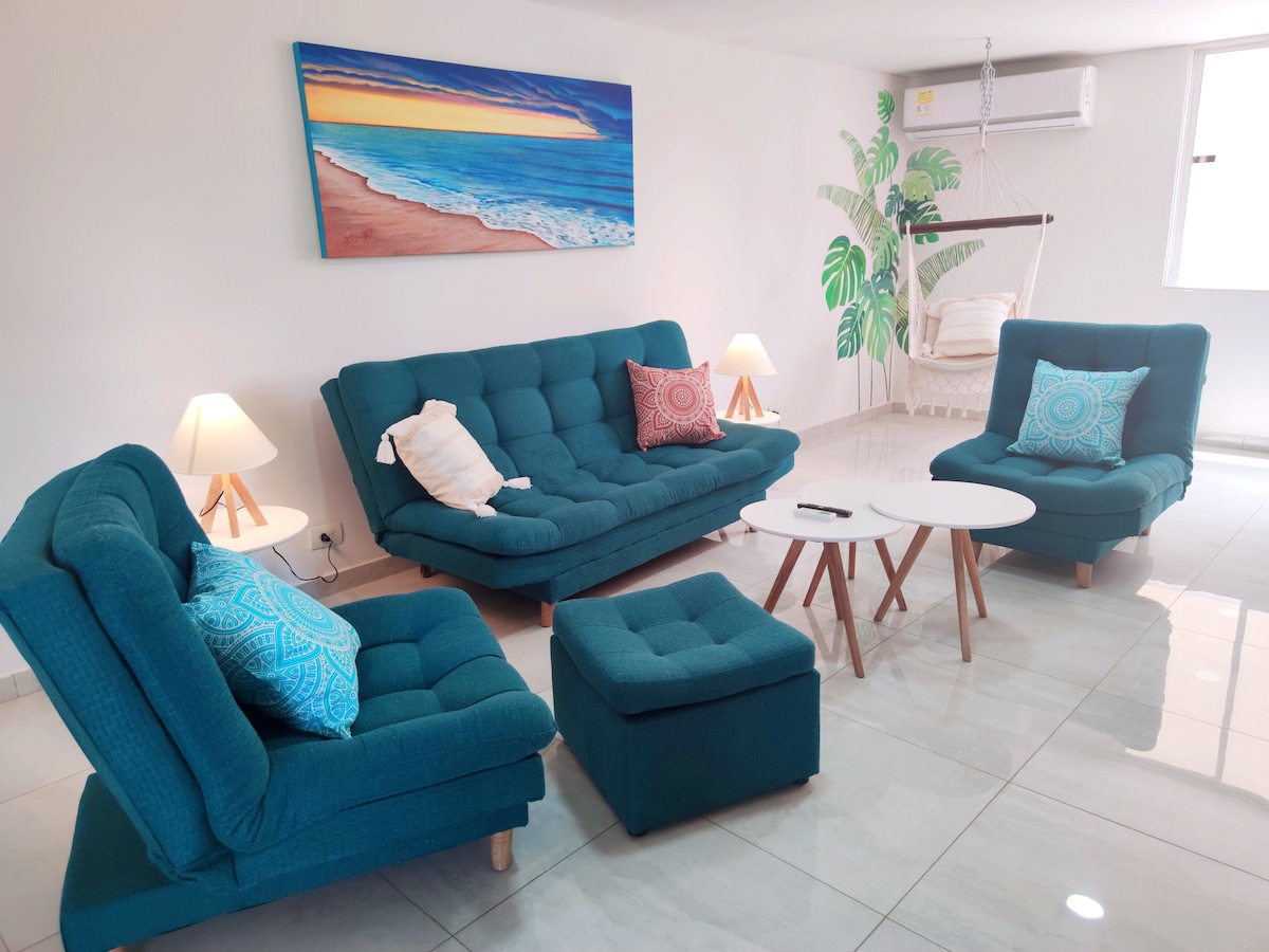A living area is presented with modern teal seating, including a spacious couch, two armchairs, and an ottoman. A light-colored coffee table occupies the center, surrounded by warm lighting from two lamps. A wall artwork depicting a beach scene adds a serene touch.
