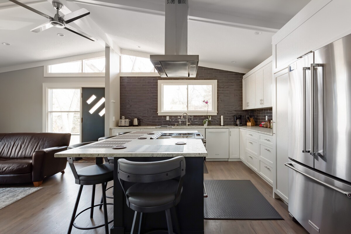 An airy kitchen features marble countertops surrounding a central island with bar seating. High windows invite natural light, illuminating gray cabinets and stainless steel appliances. A cozy leather sofa is seen in the adjacent seating area, complemented by a dark rug beneath.