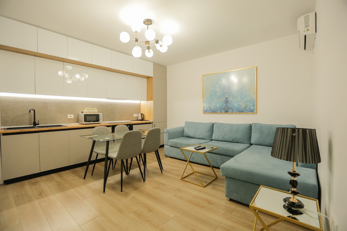 A modern living area features a light blue sofa and a sleek dining table with gray chairs. The kitchenette is equipped with essential appliances, including a microwave. A round light fixture hangs above, and an abstract painting adorns the wall, adding a touch of color.