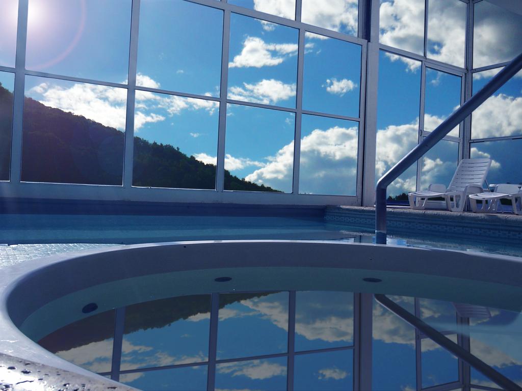 A circular jacuzzi is situated next to a large indoor pool. Expansive glass windows offer views of the blue sky and mountains. Several white lounge chairs are positioned nearby, inviting relaxation. The tranquil water reflects the surrounding scenery.