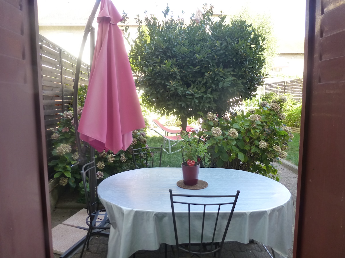 A private terrace is visible through the open doors, featuring a round table set with a small plant at the center. Four black chairs surround the table, and a pink umbrella provides shade. Lush greenery and blooming flowers frame the cozy outdoor space.