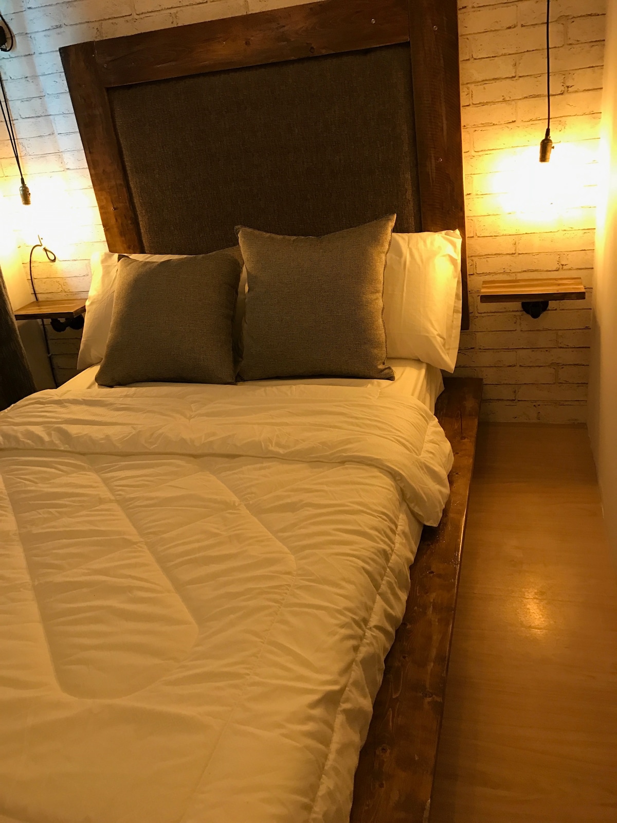 A rustic-themed sleeping area features a spacious bed with a wooden headboard and two accent pillows. Soft lighting is provided by wall-mounted fixtures on either side, illuminating the textured white brick wall behind. A smooth wooden bedside table complements the overall design.