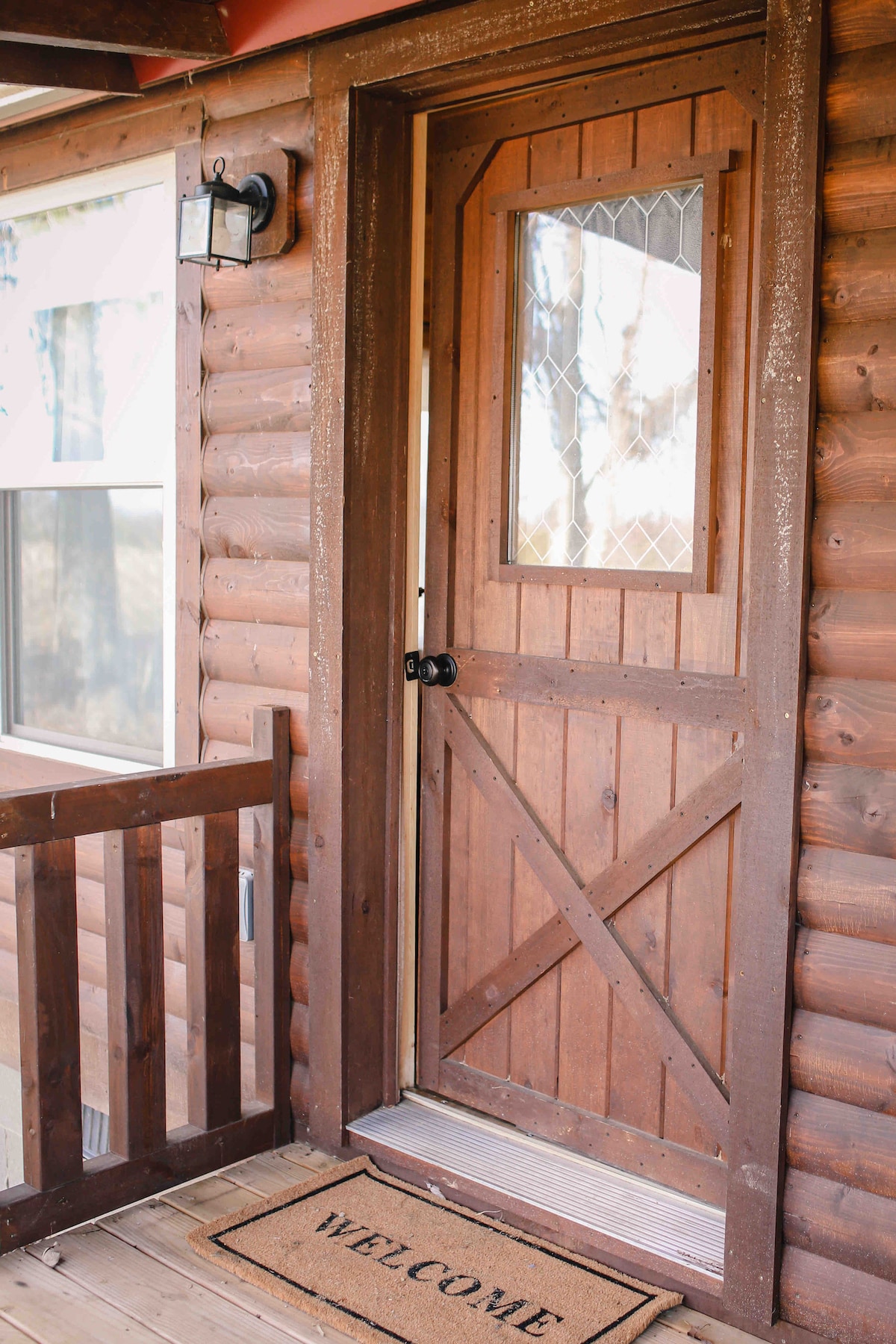 A charming wooden door with a diamond-patterned glass insert is framed by rustic logs. A 'WELCOME' doormat is placed at the threshold, inviting guests to enter. Nearby, a light fixture enhances the entrance area, while a wooden railing adds to the cabin's cozy aesthetic.
