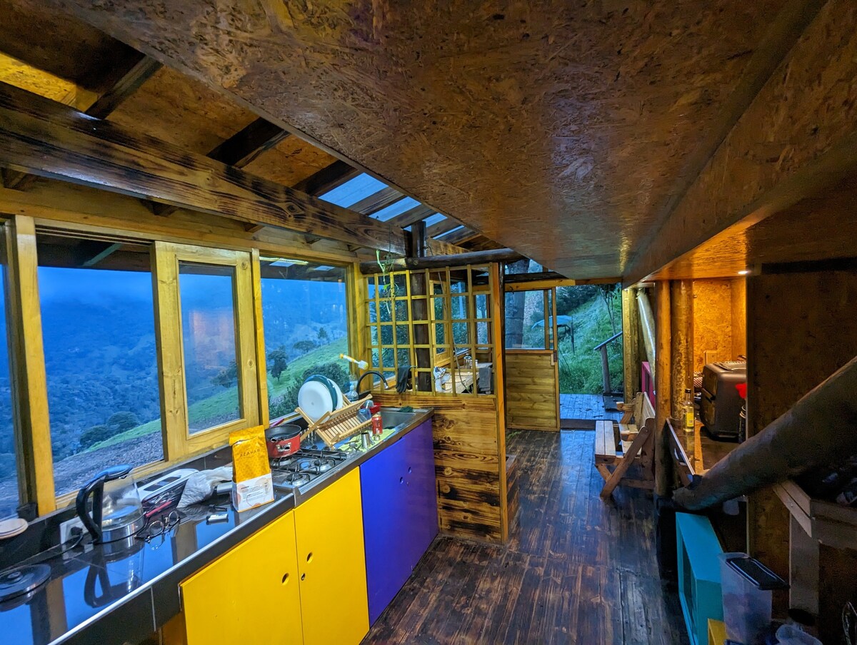The kitchen area is characterized by wooden beams and a sloped ceiling, featuring a mix of colorful cabinets. Natural light enters through large windows, illuminating the space. Cooking appliances and utensils are arranged on spacious countertops, offering an inviting environment for preparing meals.