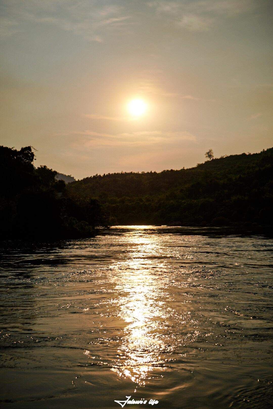 A serene river is reflected in the golden hues of the setting sun, with lush hills visible in the background. The gentle ripples on the water create a shimmering effect, emphasizing the peaceful ambiance of the natural landscape.