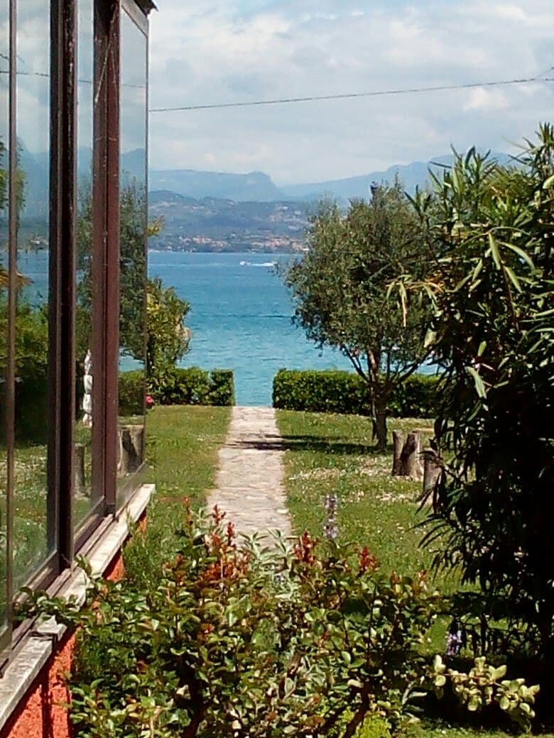 A pathway lined with greenery leads toward a scenic view of the lake, framed by low hedges and trees. The water sparkles under daylight, with distant hills visible in the background, enhancing the peaceful atmosphere of the garden.