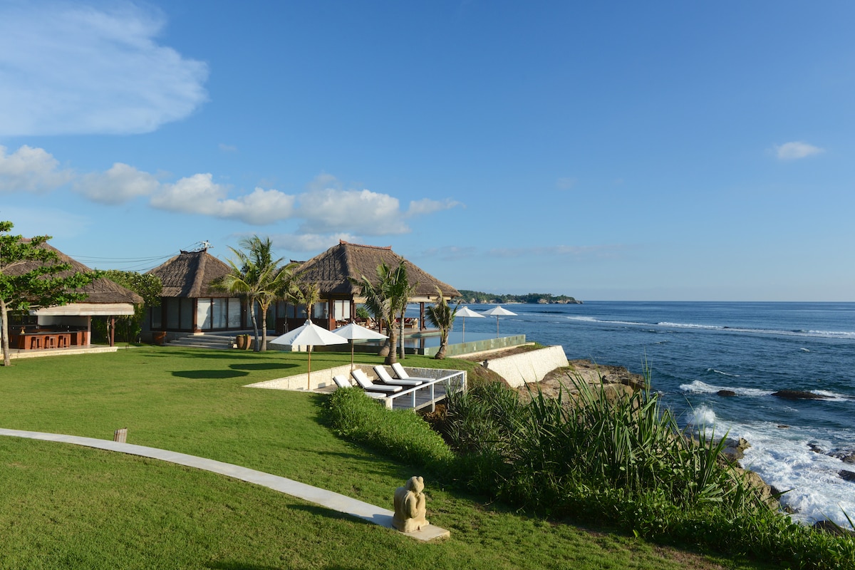 A spacious green lawn stretches towards the ocean, bordered by tropical plants. The villa features traditional thatched-roof structures, with lounge chairs arranged near the water's edge. Waves softly lap against the rocky coastline under a clear blue sky dotted with a few clouds.