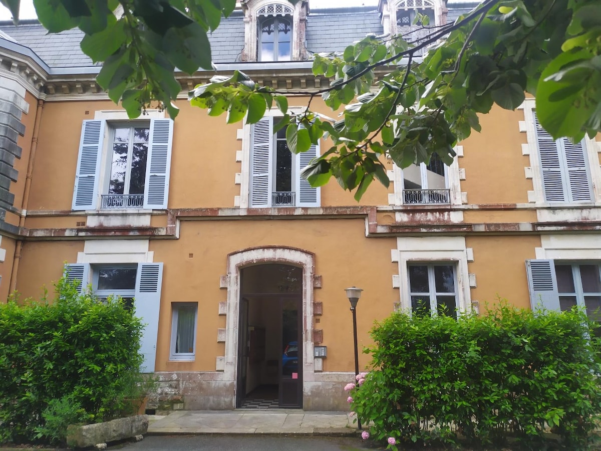 An elegant building exterior features warm-toned walls with decorative stone accents. Tall, four-paneled windows adorned with shutters are positioned symmetrically, alongside a central entrance framed by stonework. Lush greenery surrounds the entrance, complemented by flowering shrubs and a lamp post beside a pathway.