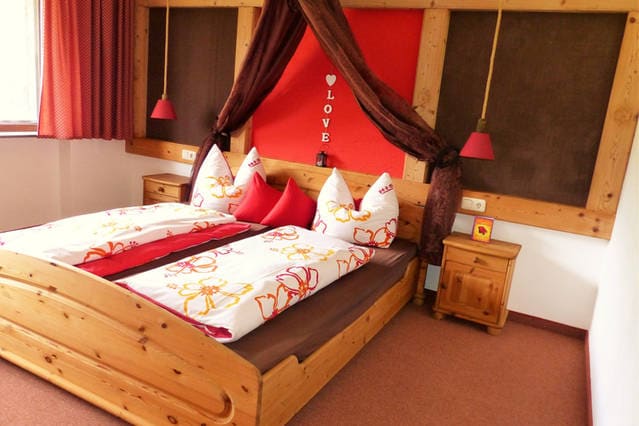 A spacious bedroom features a wooden bed adorned with decorative pillows and a floral-patterned duvet. Dark curtains frame the window, while bedside lamps provide gentle lighting. The walls display a warm color scheme, creating a cozy and inviting atmosphere.