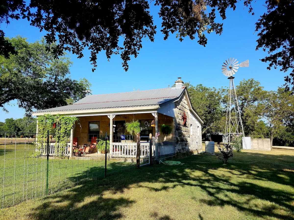 The exterior of the house is showcased with a stone façade and a metal roof. A fenced yard is visible, along with a windmill and lush greenery surrounding the home. The porch features a seating area, offering a welcoming outdoor space.