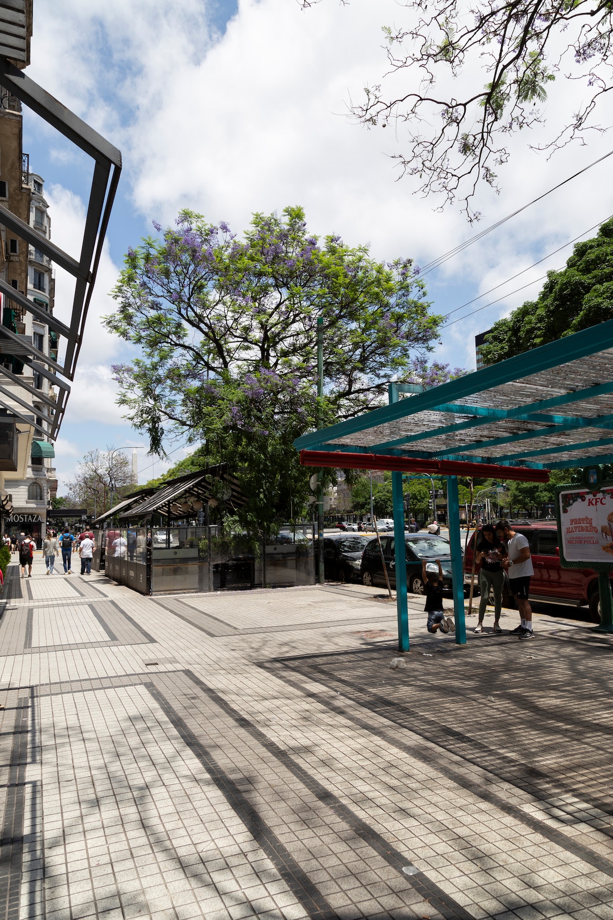 A lively city street is depicted, featuring a vibrant blue shelter at a bus stop. Nearby, a large blossoming tree provides shade. The scene includes pedestrians and parked cars, with light filtering through the leaves and creating dynamic shadows on the tiled walkway.