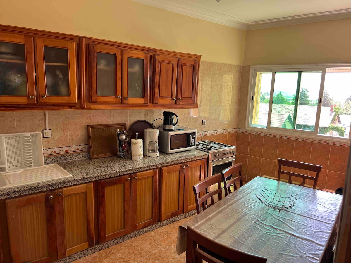 A well-equipped kitchen features wooden cabinets and a spacious countertop. Kitchen appliances, including a coffee maker and microwave, are visible. A dining table with multiple chairs sits nearby, and large windows allow natural light to fill the space, offering views of the outside.