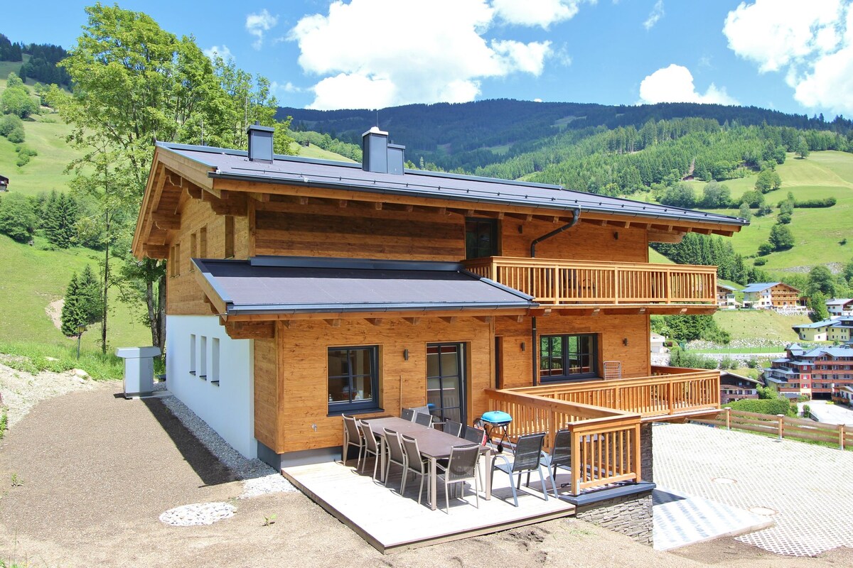 The chalet's exterior features a blend of natural wood and modern design, showcasing multiple balconies and a spacious terrace. Surrounding hills are visible in the background, with a clear blue sky above. Ample outdoor seating is provided, suitable for gatherings.