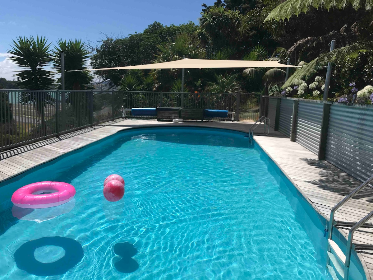 A refreshing pool is surrounded by landscaped gardens and features a wooden deck. A shaded area provides rest space. Brightly colored floats are visible in the clear water, enhancing the inviting appearance of the pool area.