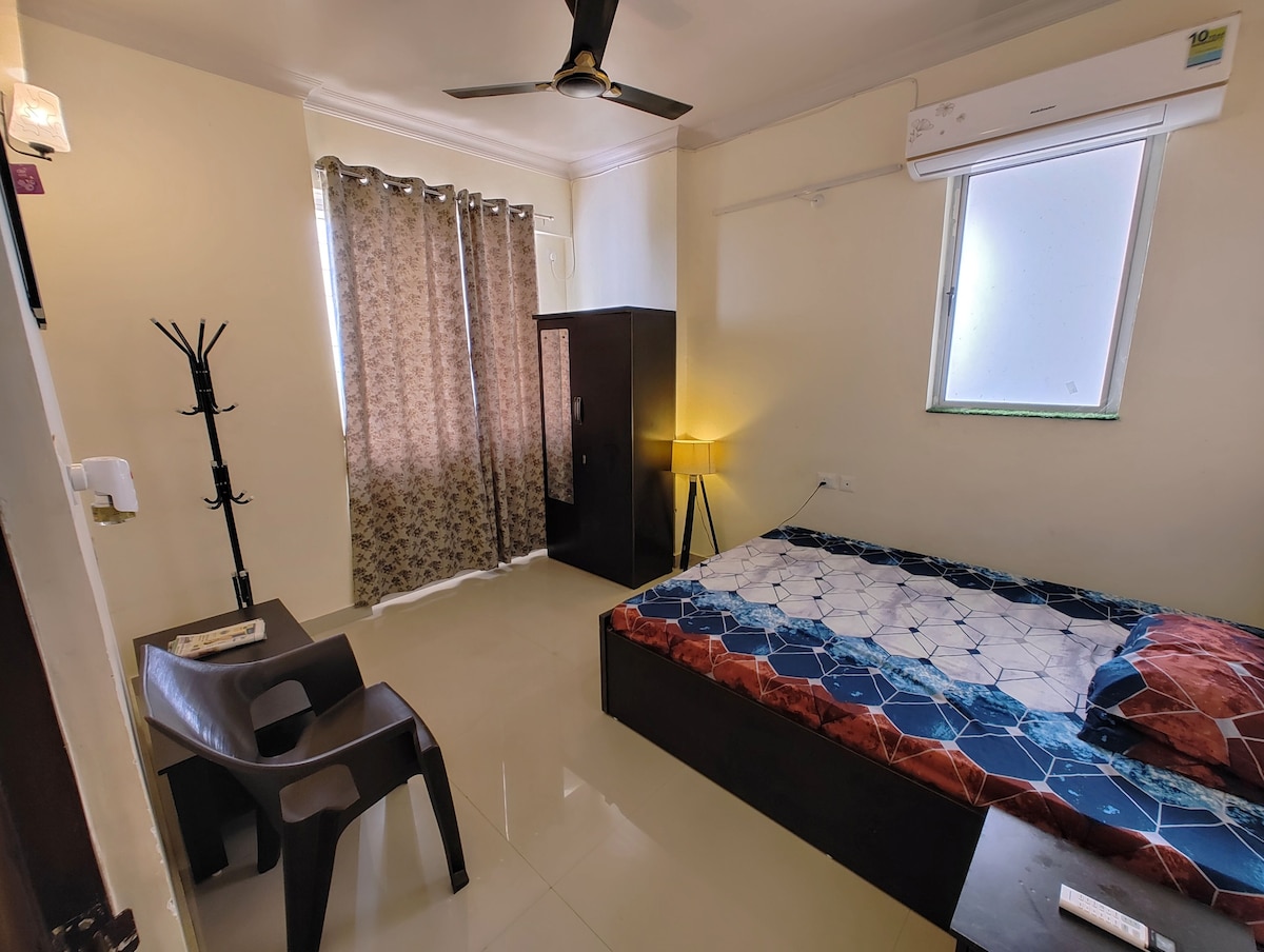 A well-lit bedroom features a spacious bed with a blue and white patterned bedspread. Two chairs are positioned next to a small table, and a wardrobe stands against the wall. Curtains cover the window, allowing soft light to filter through.