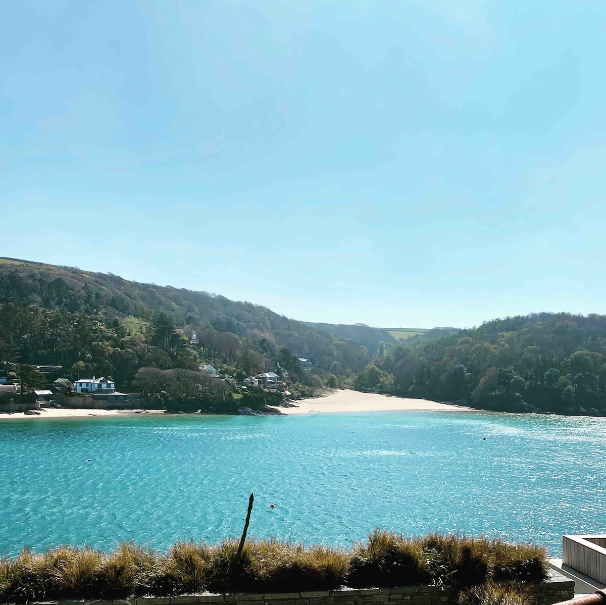 A serene coastal view captures a tranquil bay with clear turquoise waters. Lush green hills frame the beach, while a sandy shore is visible in the distance under a clear blue sky.
