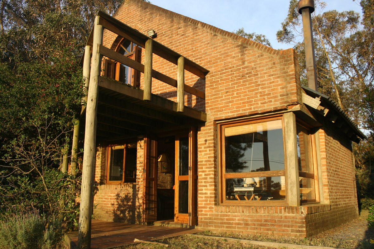 A brick house features large windows and a wooden deck, allowing for natural light to fill the interior. The upper balcony overlooks the surrounding greenery, and a chimney is visible on the side. The entrance is framed by natural wooden elements, integrating the structure with its environment.