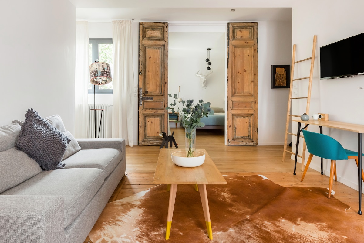 A light-filled living area is featured, showcasing a grey sofa beside a wooden coffee table atop a brown area rug. Vintage double doors lead to a serene bedroom, while a wall-mounted TV and a small desk add functional touches to the space.