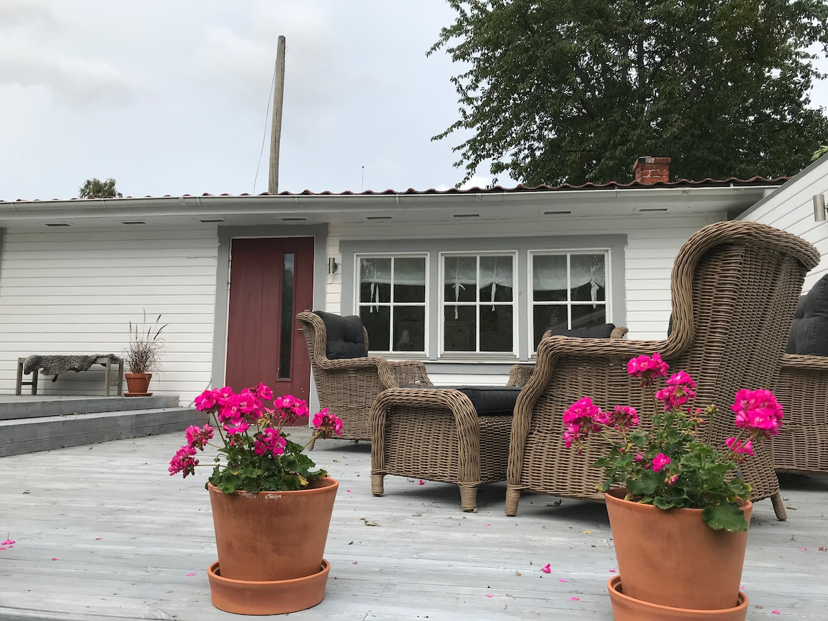 A charming outdoor seating area is presented with comfortable wicker chairs positioned on a wooden deck. Potted pink flowers add a vibrant touch, while the entrance showcases a double door framed by large windows. A serene atmosphere is created by the surrounding greenery.