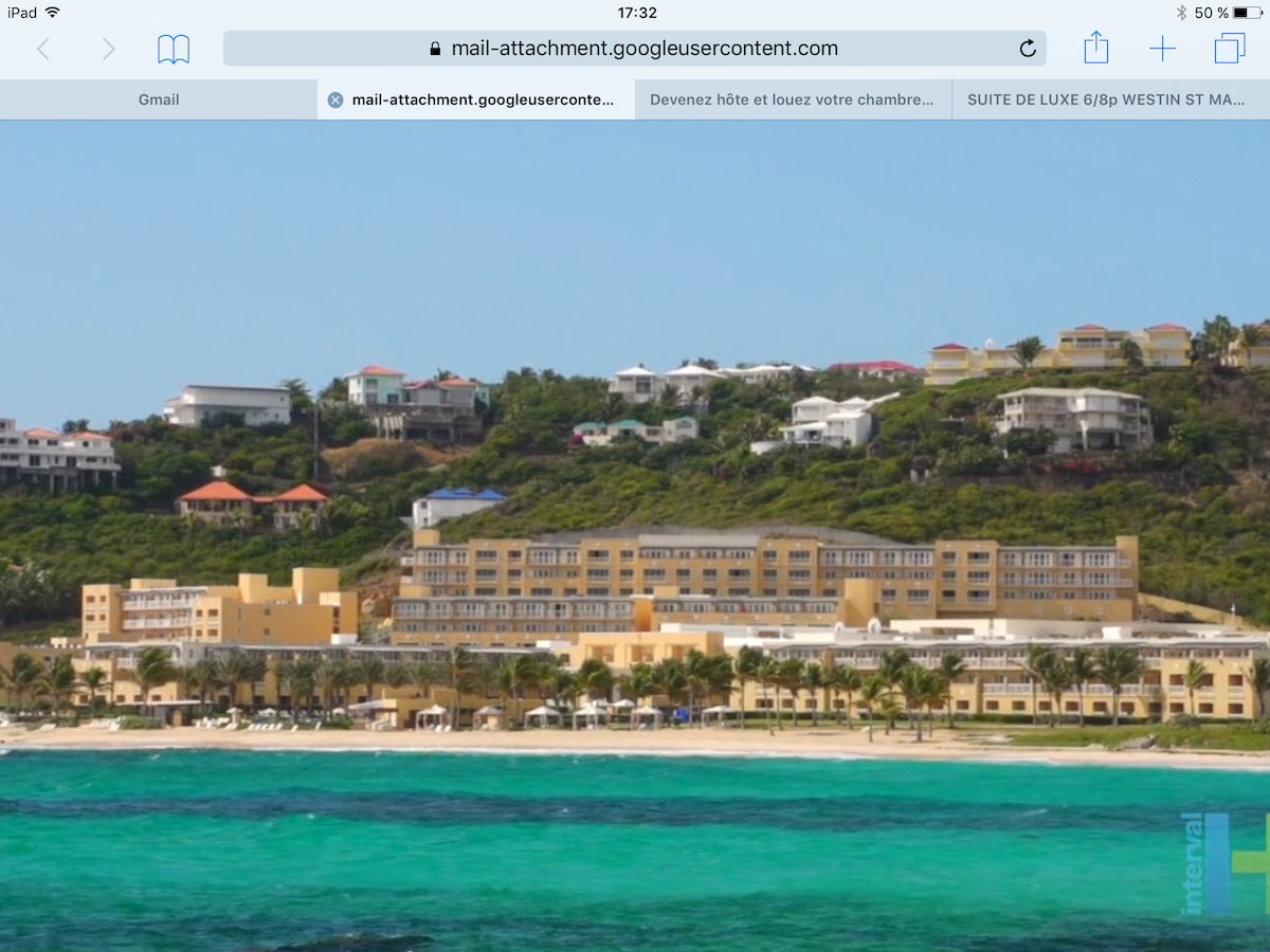 A beachfront view showcases a sprawling resort against a backdrop of lush green hills. The building's modern architecture is highlighted, with multiple levels and large windows. Pristine turquoise water and sandy beach create a serene coastal setting, complemented by nearby residential structures on the hillside.