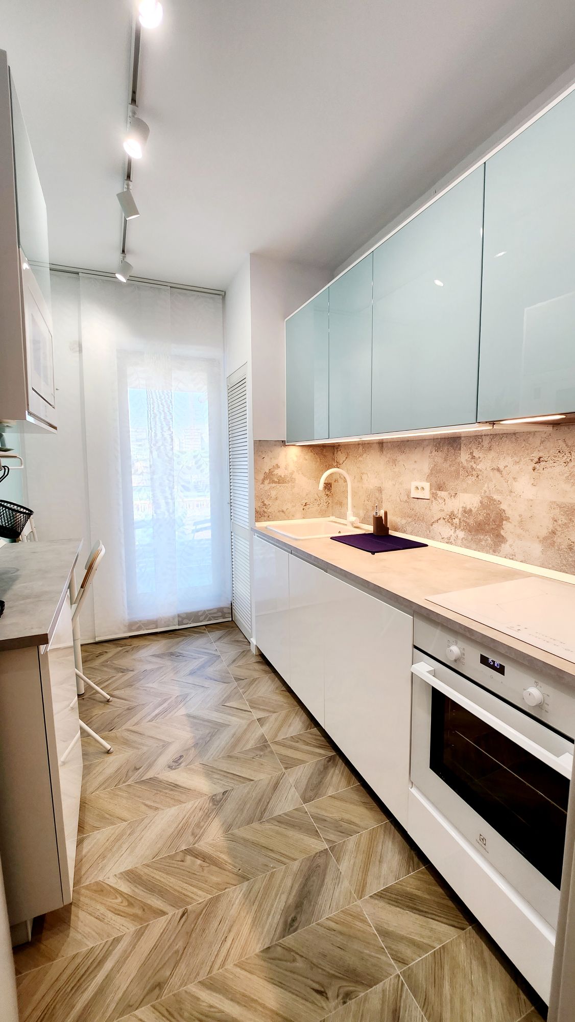 A modern kitchen features smooth white cabinetry with contrasting light blue upper cabinets. An oven and stove are positioned under a sleek countertop. Natural light enters through a nearby window, illuminating the herringbone-patterned flooring and creating a spacious cooking area.