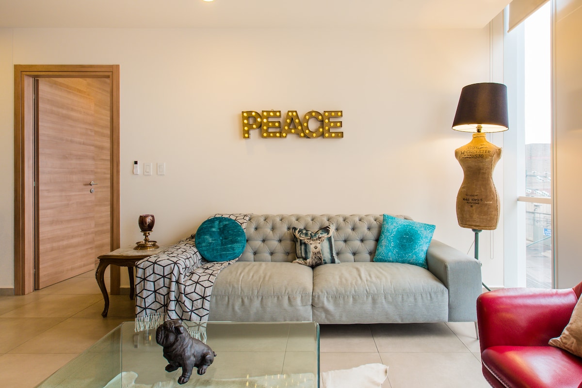 A light-filled living space features a tufted grey sofa adorned with teal cushions and a decorative throw. An illuminated 'PEACE' sign hangs on the wall, complemented by a stylish floor lamp. A glass coffee table sits in front, displaying a small decorative figurine.
