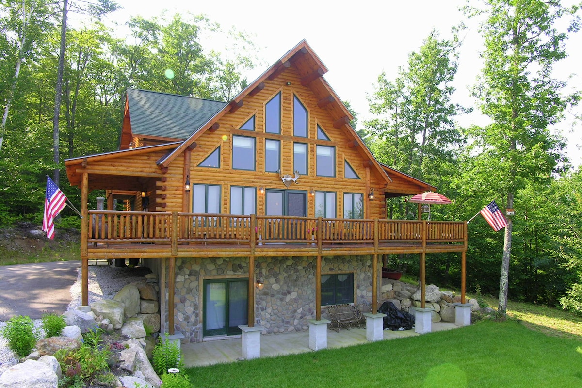 The log cabin features a spacious facade with a large deck adorned by American flags. Floor-to-ceiling windows showcase the natural surroundings, while the stone base provides a solid foundation. Lush greenery and a well-maintained yard enhance the inviting outdoor space.
