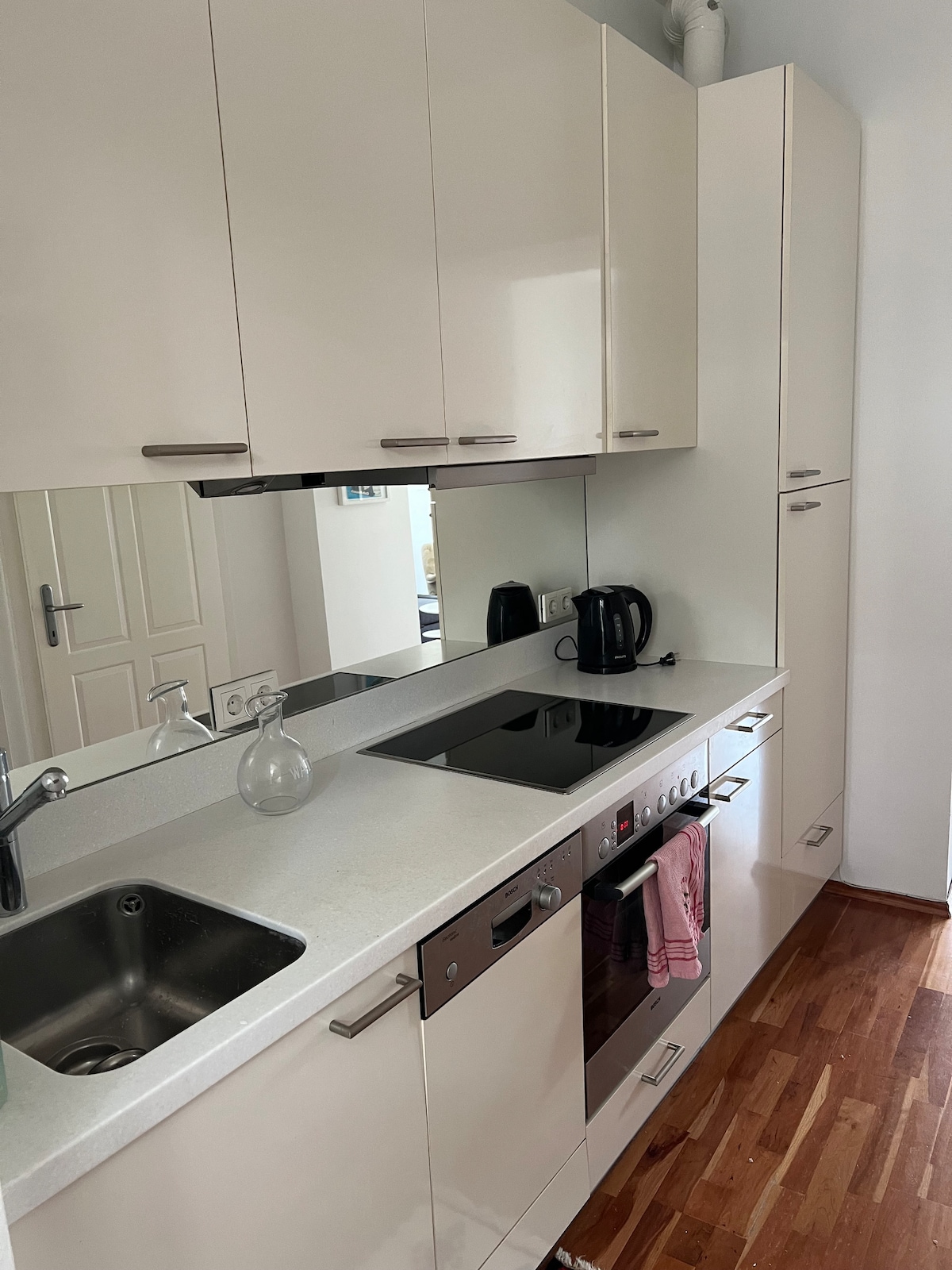 A modern kitchen is displayed with sleek white cabinetry and a glossy countertop. Essential appliances, including a kettle and an integrated oven, are visible. A mirrored backsplash reflects the organized space, which features a stainless steel sink and a ceramic cooktop, enhancing functionality.