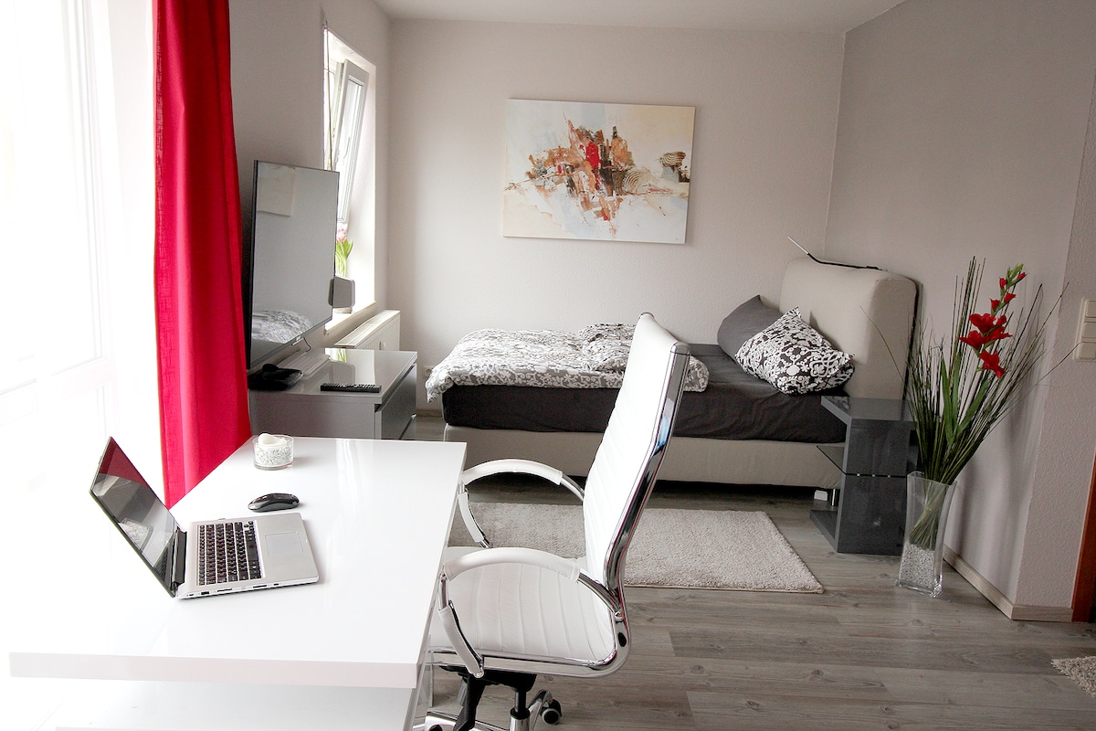 A modern bedroom workspace features a spacious desk paired with a sleek white chair. A comfortable bed is positioned against the wall, adorned with patterned bedding. Natural light brightens the room through large windows, complemented by decorative plants and a wall-mounted television.
