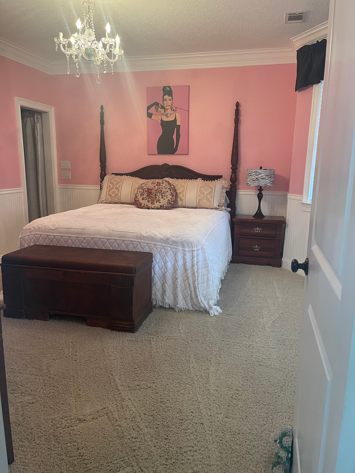 A bedroom features a spacious bed with a white quilt atop a dark wooden frame. A decorative light fixture hangs from the ceiling, and a pink wall complements the room's aesthetic. A nightstand with a lamp and a bench at the foot of the bed are also visible.