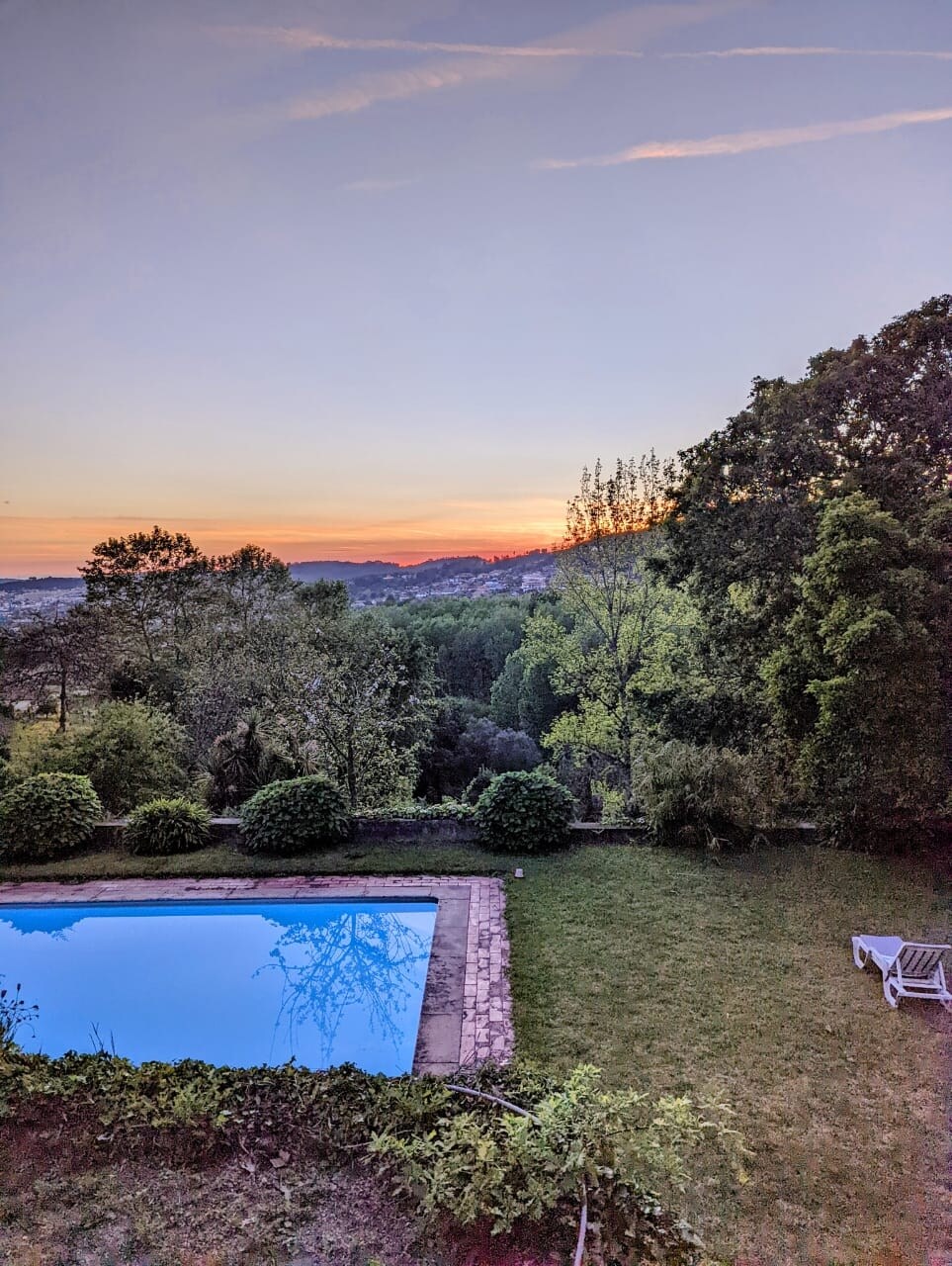 A tranquil outdoor scene features a swimming pool reflecting the soft colors of a sunset. Lush greenery surrounds the pool area, with trees and bushes creating a serene landscape. The horizon displays warm hues as the day transitions to evening.