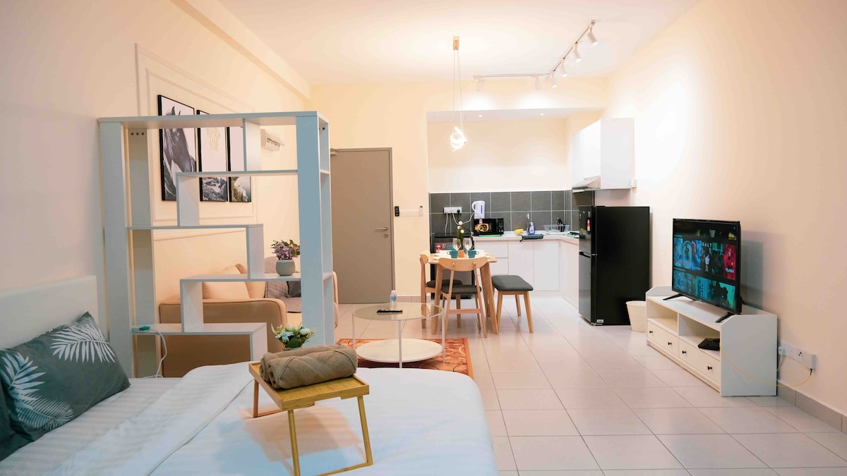 The studio features a cozy layout with a queen size bed, a dining area for two, and a well-equipped kitchenette. Natural light illuminates the space, revealing a modern living area with a TV and comfortable furnishings. Decorative plants add a touch of greenery.
