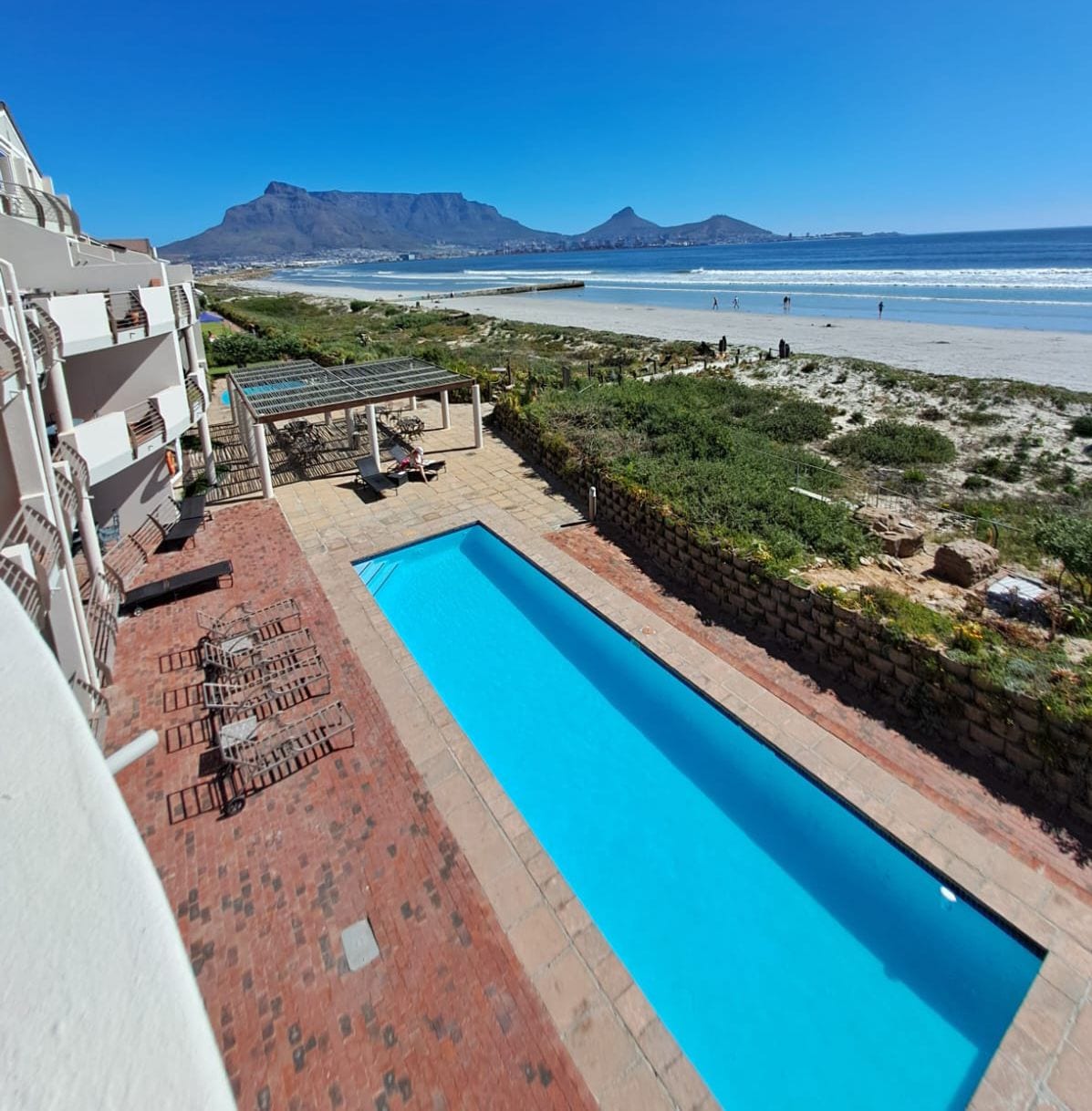 An aerial view of a long swimming pool, bordered by a brick patio and loungers. The beach and ocean are visible in the background, along with Table Mountain against a clear blue sky. Lush greenery adds a natural element to the scene.