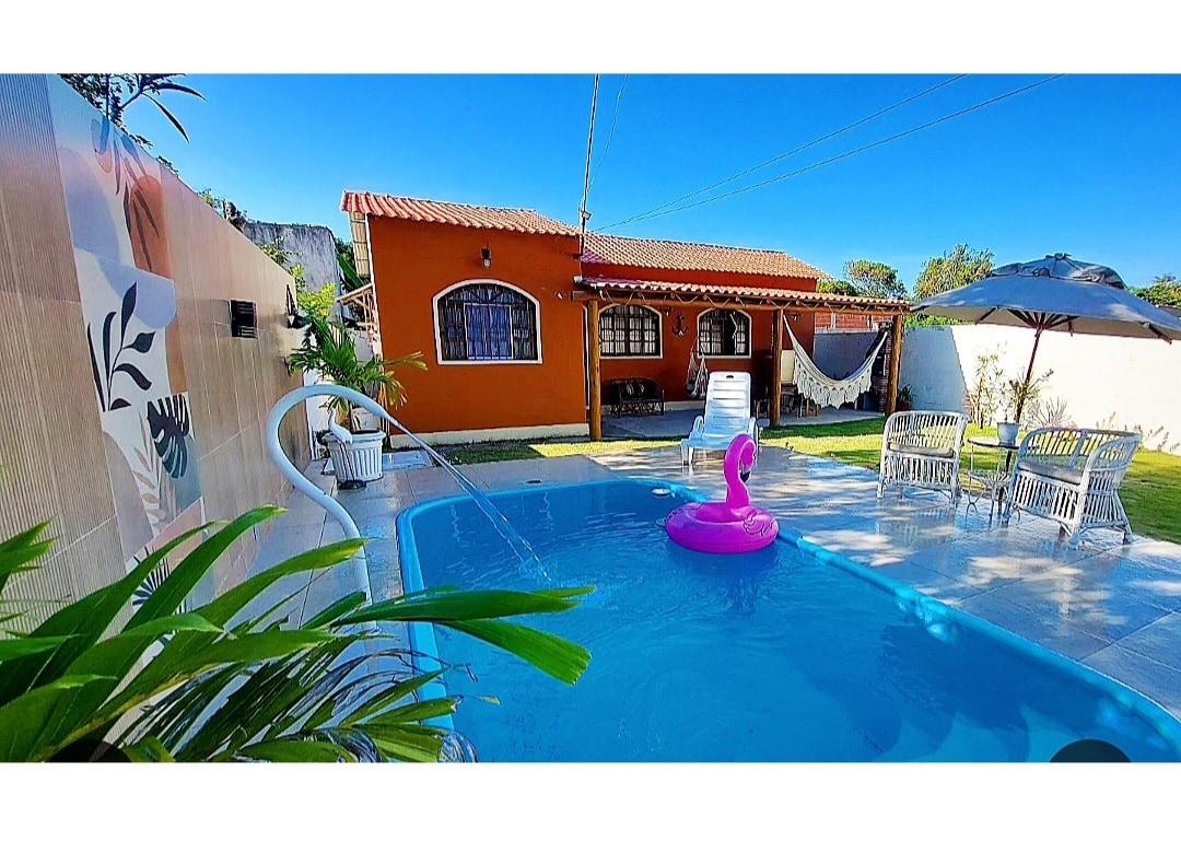 A refreshing swimming pool is featured with a pink inflatable flamingo floating on the water. Surrounding the pool are comfortable lounge chairs and a sun umbrella. In the background, a cozy orange house is visible, complemented by green grass and tropical plants.