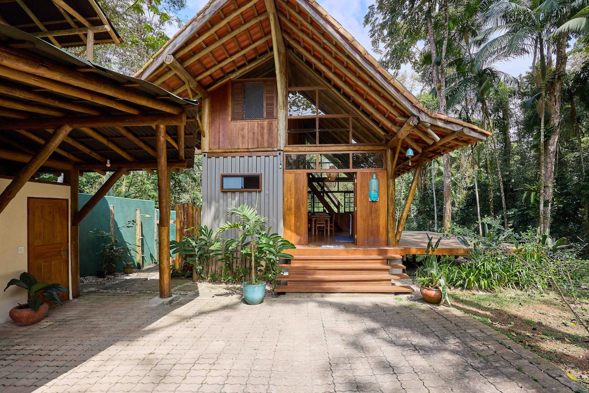 The exterior view of the bungalow showcases a wooden structure with a sloped roof and large windows. Surrounding greenery emphasizes the natural setting. A spacious patio leads to the entrance, and a driveway provides access to the garage area.