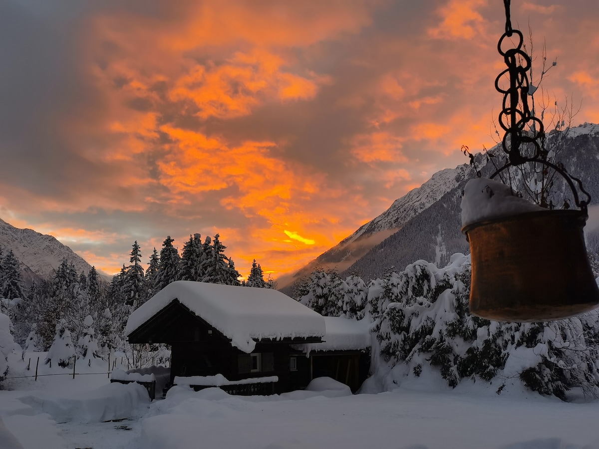 A cozy chalet is pictured amid a snowy landscape, with a colorful sunset sky casting warm hues over the scene. The surrounding mountains and trees are blanketed in snow, while a rustic bell hangs in the foreground, adding charm to this tranquil setting.